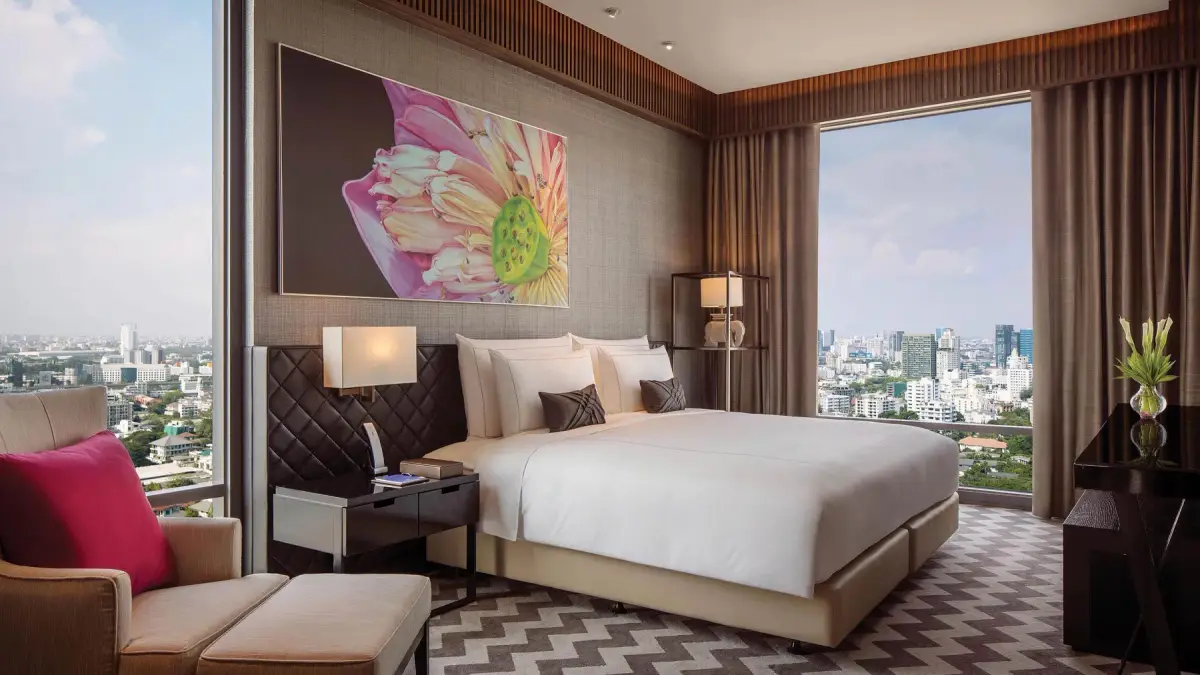 A bedroom with a quilted headboard, a lotus painting on the wall and a city view through glass