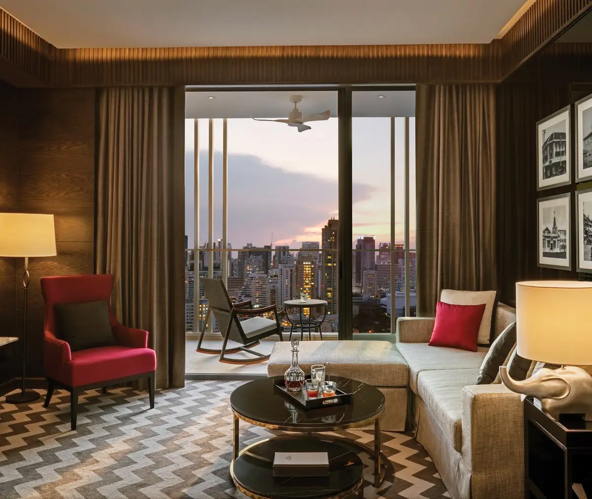 A sitting area with a lamp, a red armchair and a balcony looking over the city at dusk