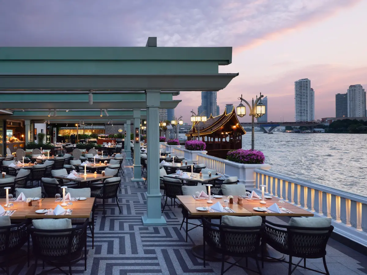 Tables set for dinner on a covered riverside terrace with the Bangkok skyline beyond