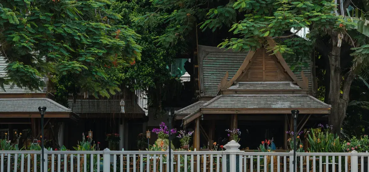 Two teak pavilions with steep tiled roofs among tropical planting beside a white fence