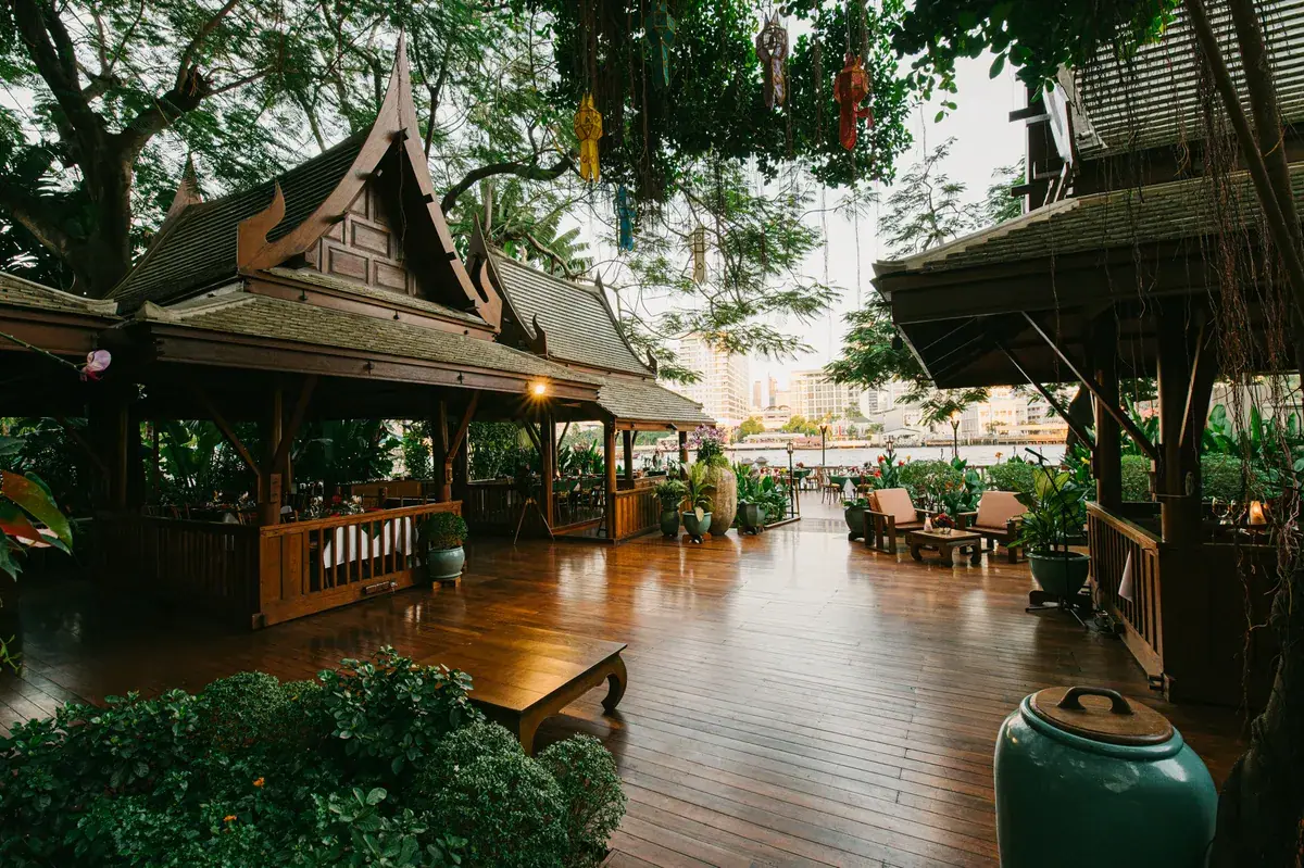 A teak Thai pavilion with a tiled roof standing in a garden at the river's edge