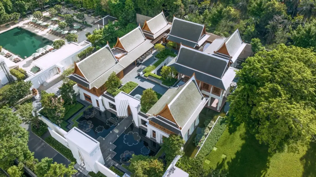 An aerial view of low white pavilions with steep tiled roofs set among lawns and trees