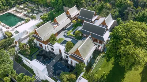 An aerial view of low white pavilions with steep tiled roofs set among lawns and trees