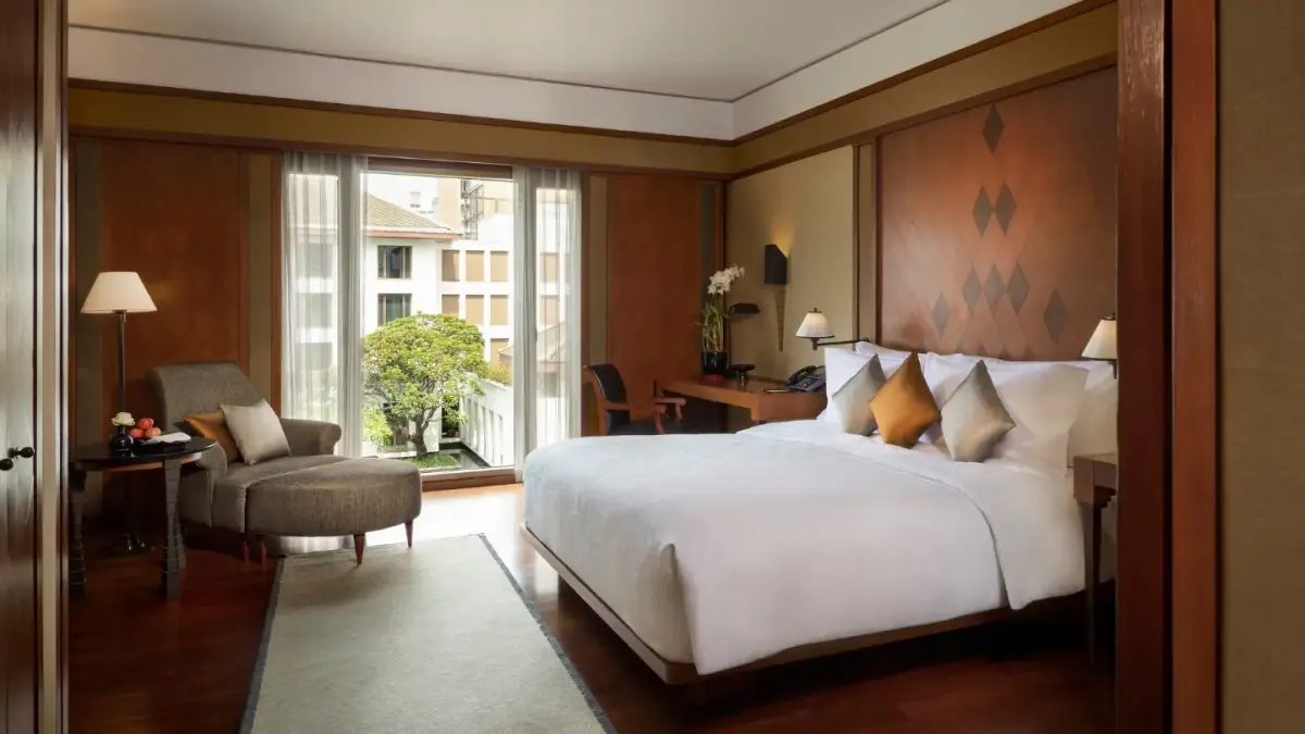 A bedroom in teak and cream with a balcony and green trees filling the window
