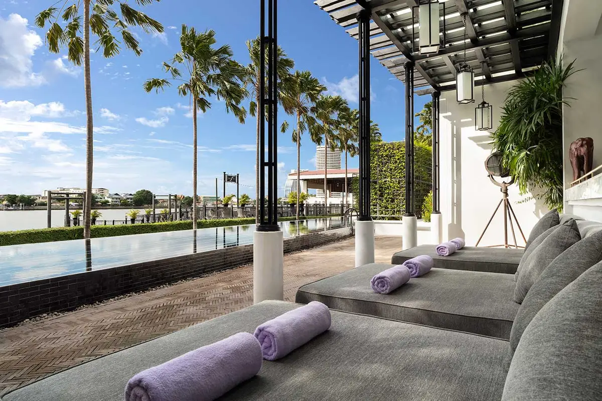 A long pool and a colonnaded walkway with sun loungers and palms beside the river