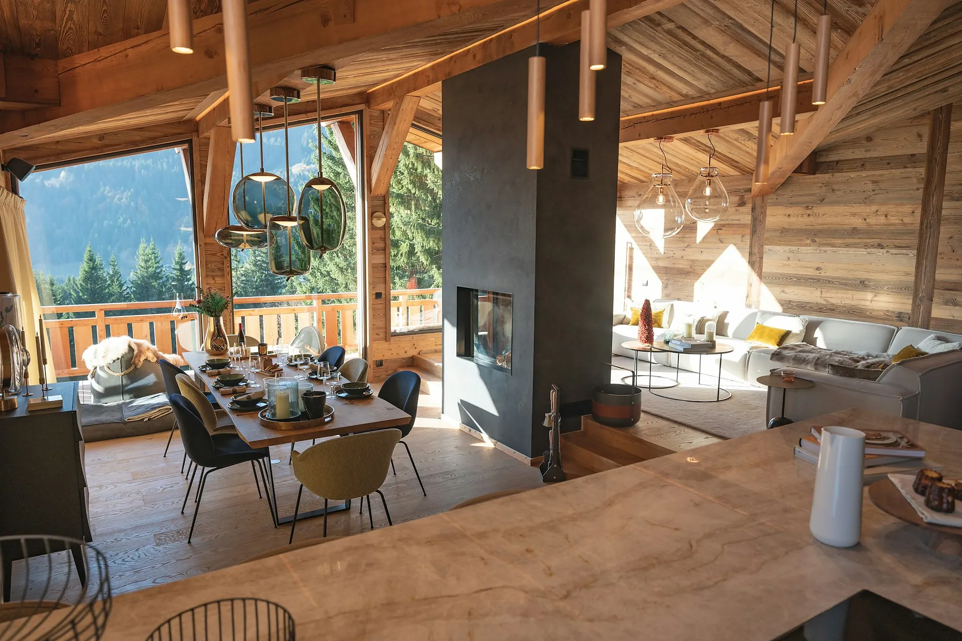 A timber chalet interior with a long dining table, an open fire and mountain windows