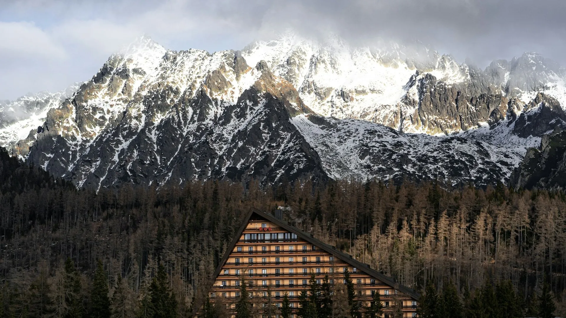 A modern timber hotel standing among dark pines beneath a long wall of snow covered peaks