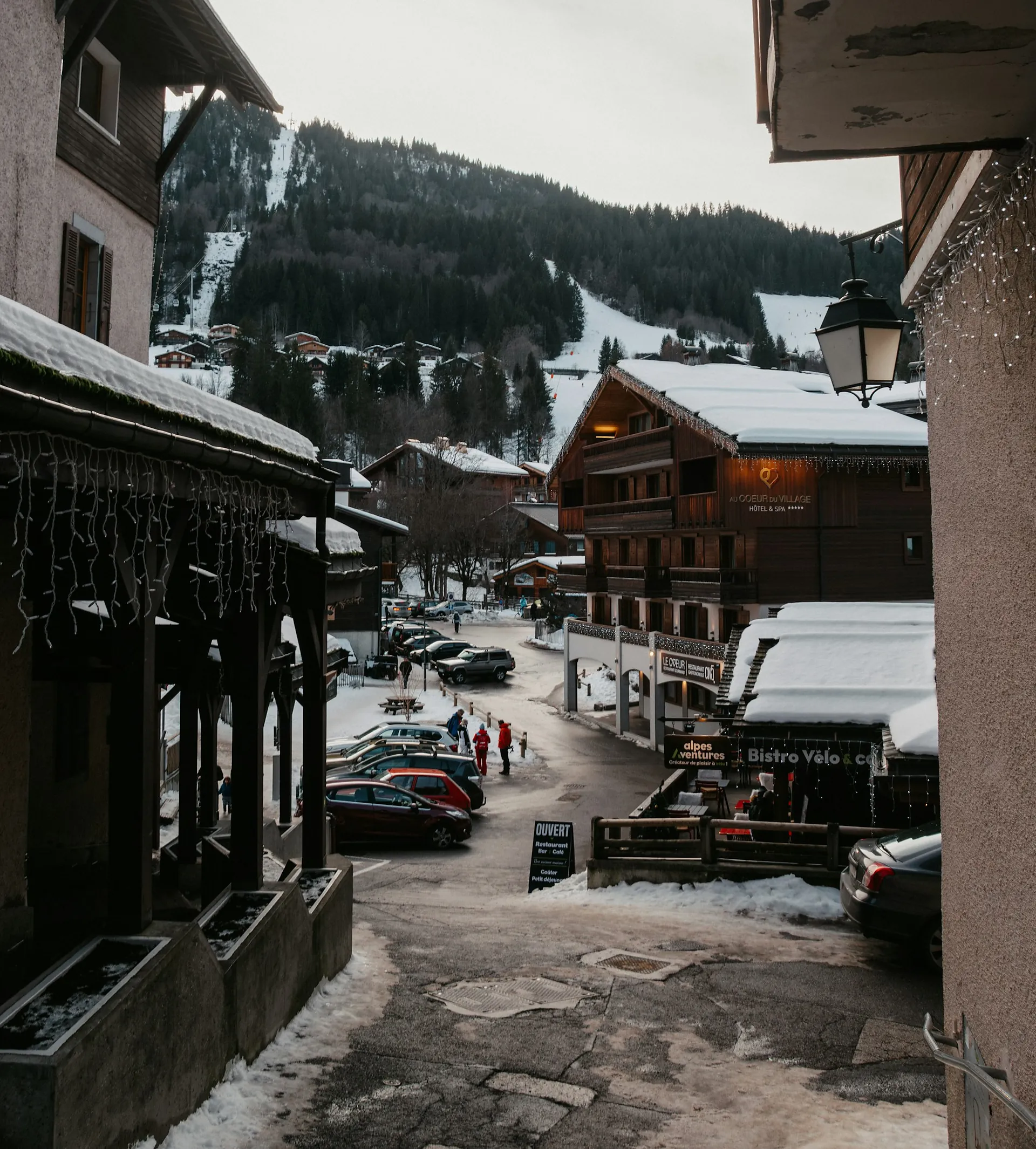A snowy village street of timber buildings with pisted slopes rising behind