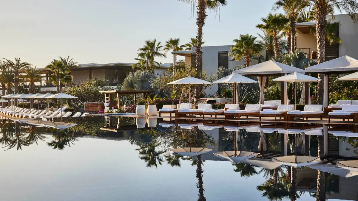 A long still pool lined with cabanas and palms reflecting the sky