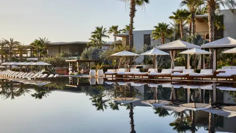 A long still pool lined with cabanas and palms reflecting the sky
