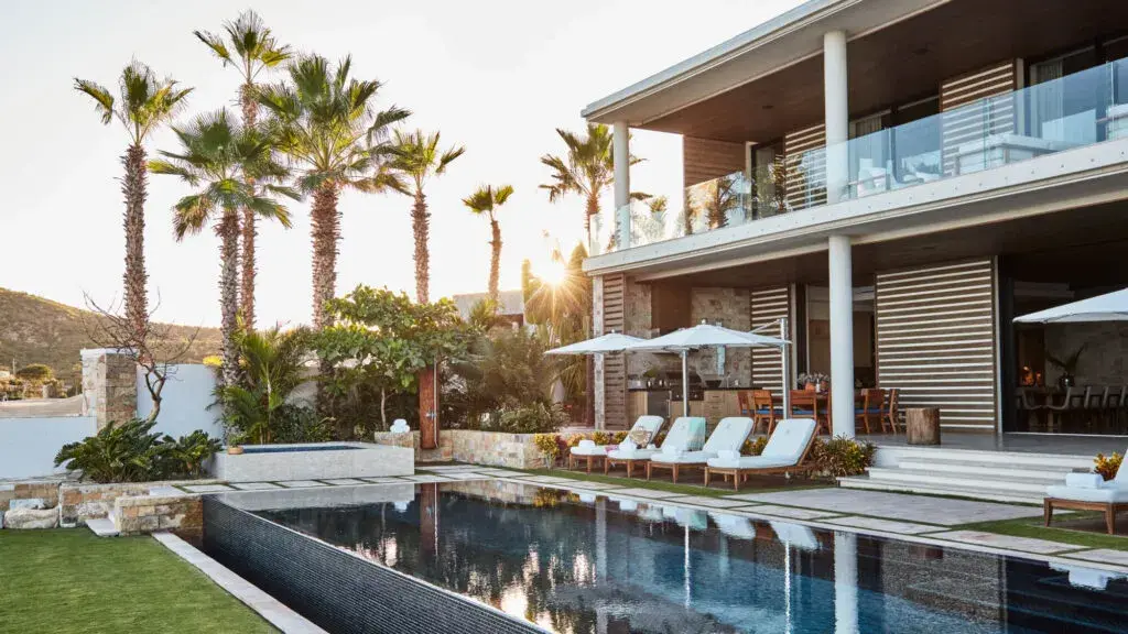 A two storey villa with a long dark pool, loungers and tall palms in evening light