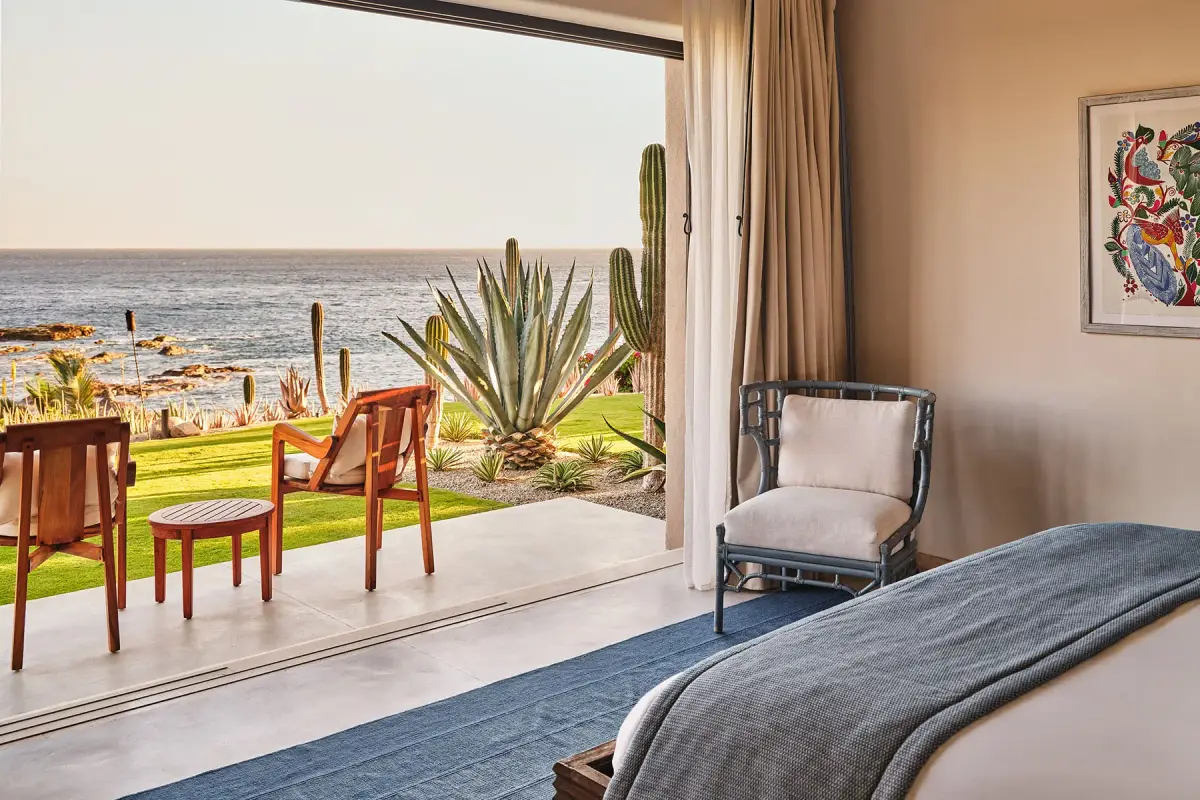 A bedroom with doors folded back onto a lawn, cactus planting and the sea