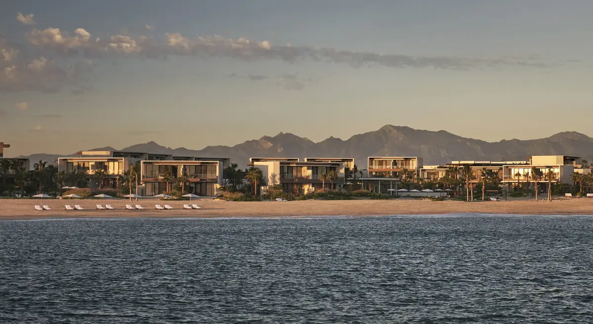 A long low resort along a flat beach with mountains rising behind it, seen from the water