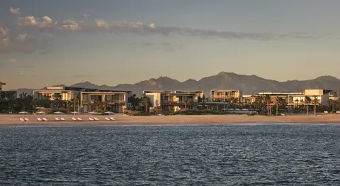 A long low resort along a flat beach with mountains rising behind it, seen from the water