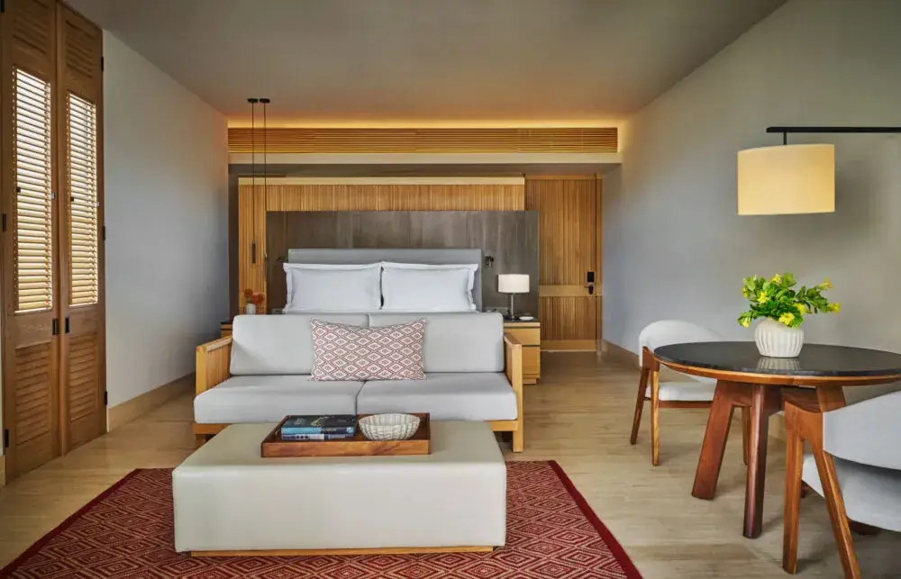 A pale bedroom with a timber headboard, a round table and a view through to the terrace