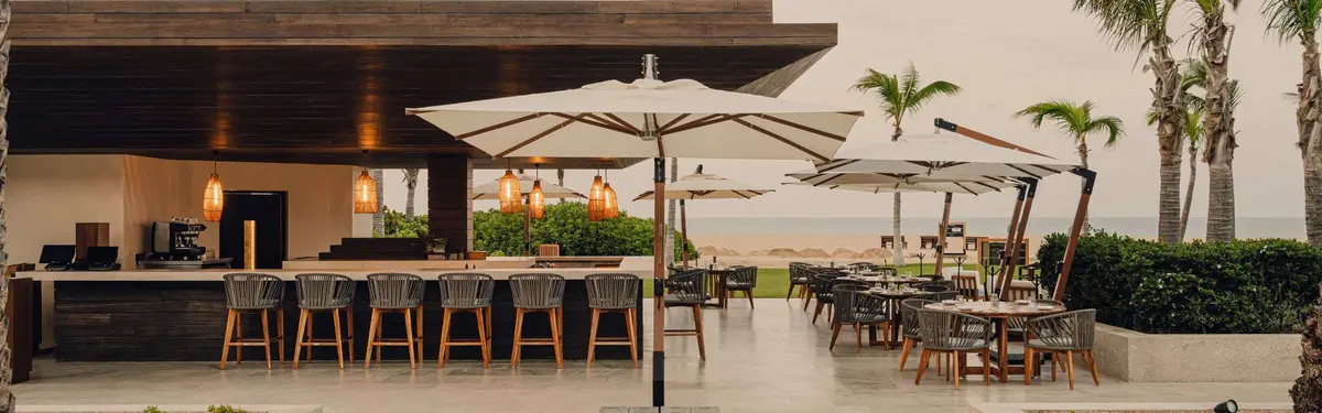A bar counter with high stools under cream parasols looking out to the beach
