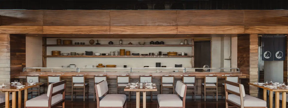 A sushi counter with pale stools and shelves of ceramics behind it