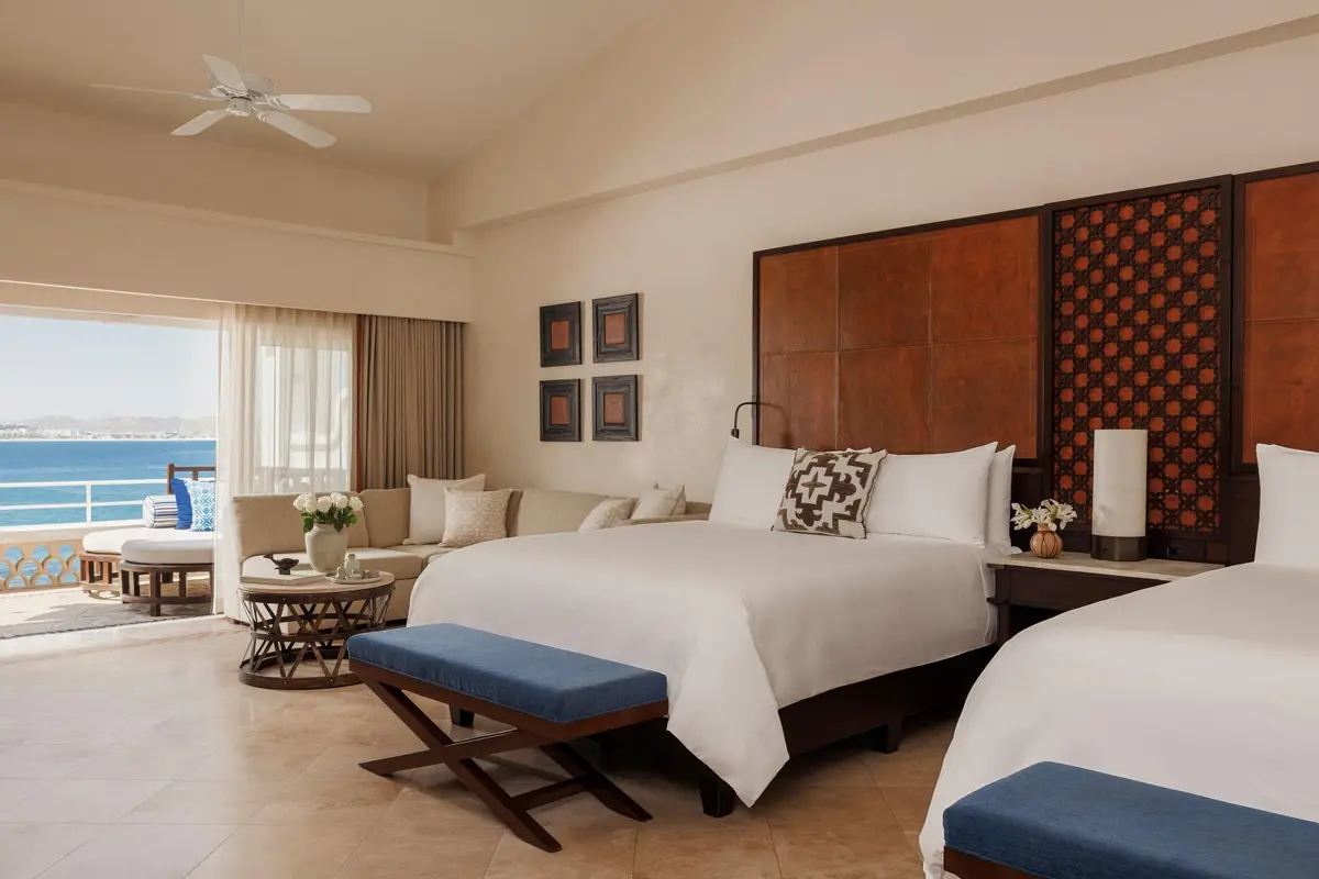 A bedroom with two beds, a dark carved headboard and doors open to the sea