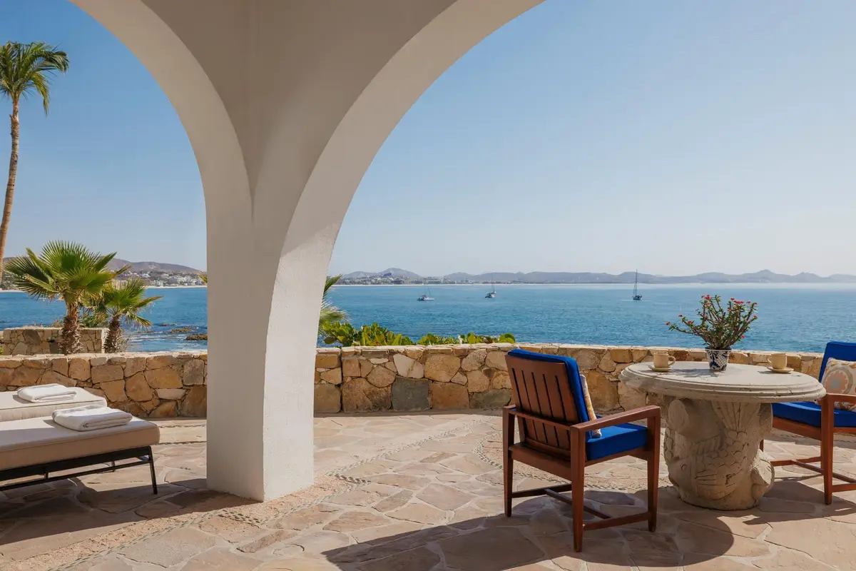 A stone terrace under white arches with daybeds and blue chairs looking over the bay