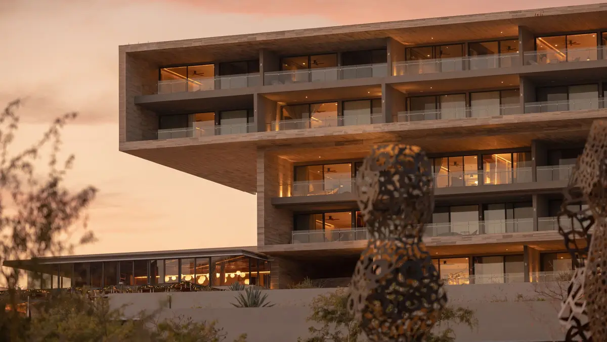 A concrete and timber facade of stacked balconies lit warm at dusk with a sculpture in front