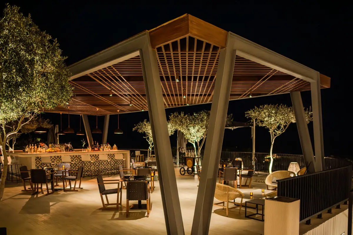 A timber pavilion restaurant lit at night with olive trees growing through the terrace