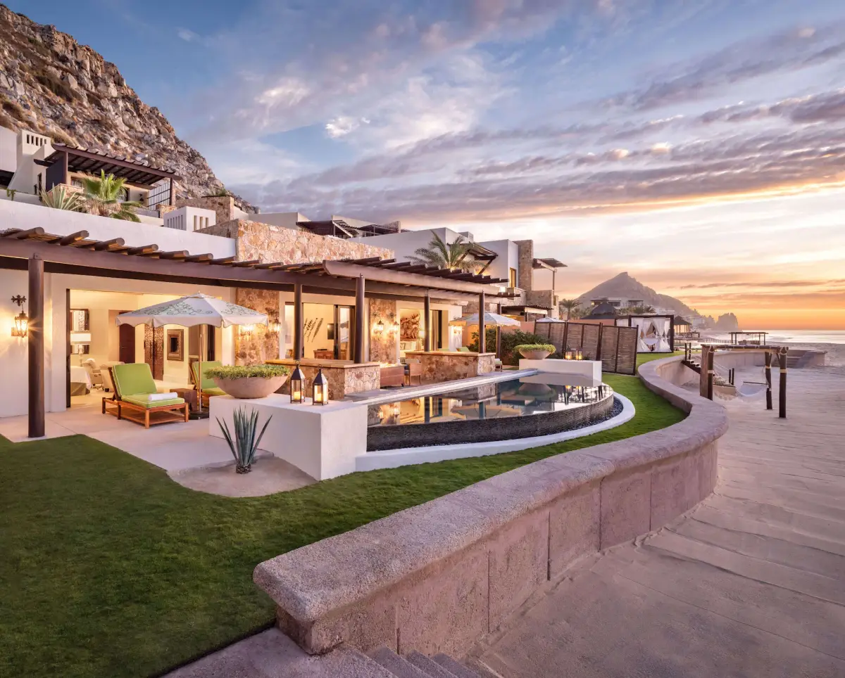 A single storey beachfront suite with a lit plunge pool and the sea behind it at dusk