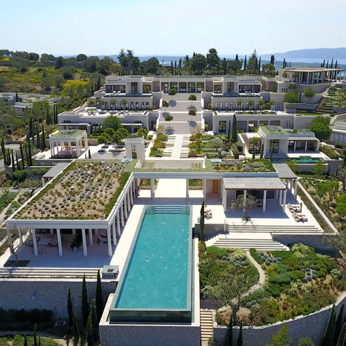 An aerial view of white pavilions and a long pool at a hilltop resort above the sea