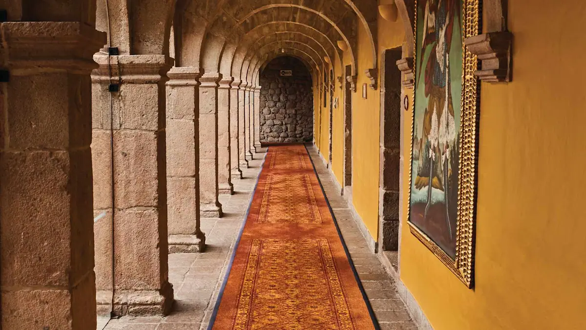 A stone cloister corridor with painted canvases along one wall and a red runner