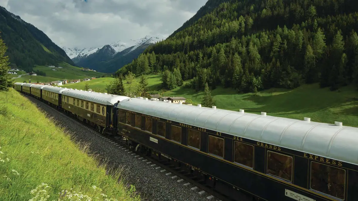 A dark blue and gold train running through a green alpine valley below snow