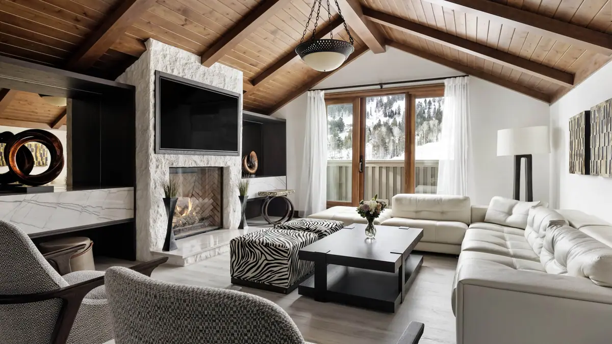 A lodge sitting room with a stone fireplace, timber beams and windows onto mountains