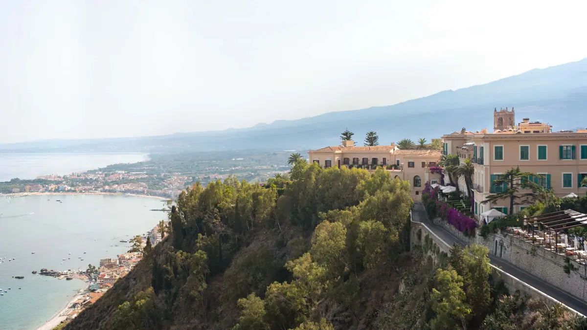A cliff above a blue bay with a terracotta coloured hotel and gardens on top of it