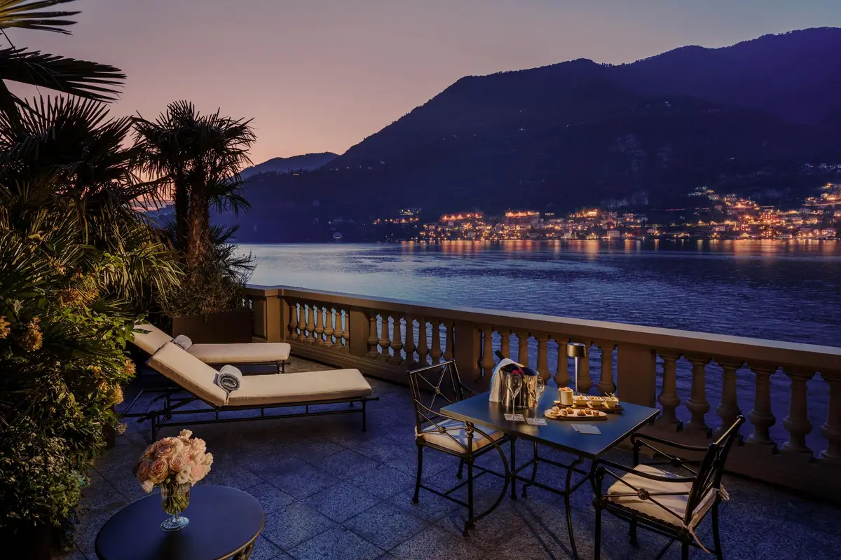 A stone terrace with loungers looking down a lake towards lit towns at dusk
