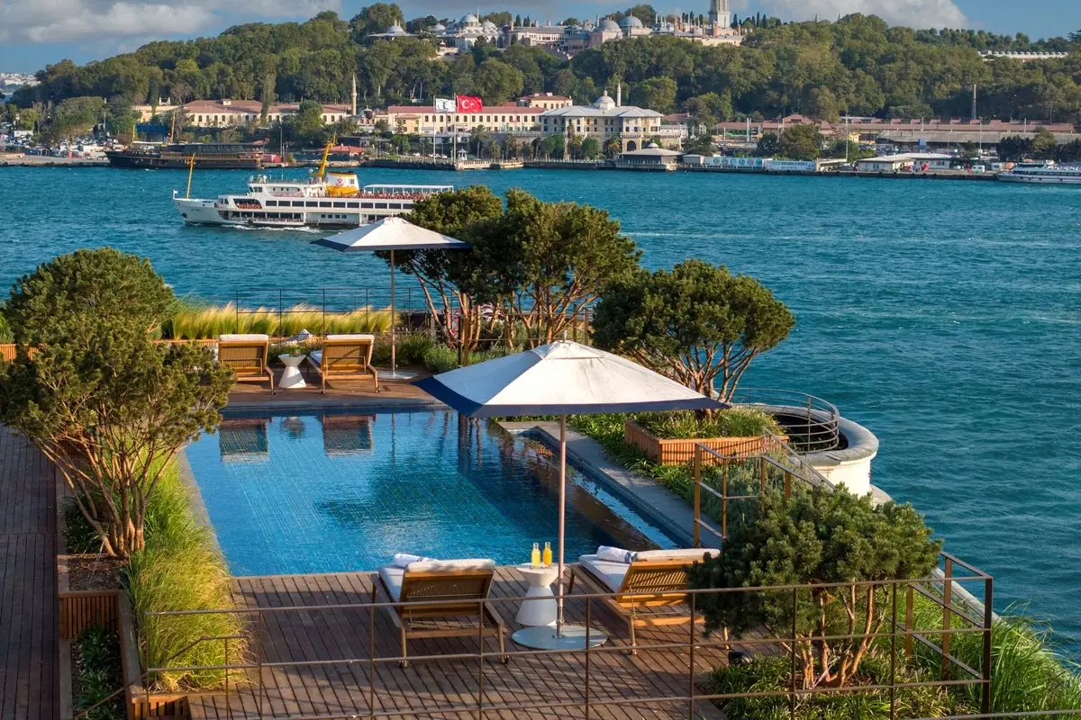 A pool deck with umbrellas and loungers beside a wide strait with ferries on it