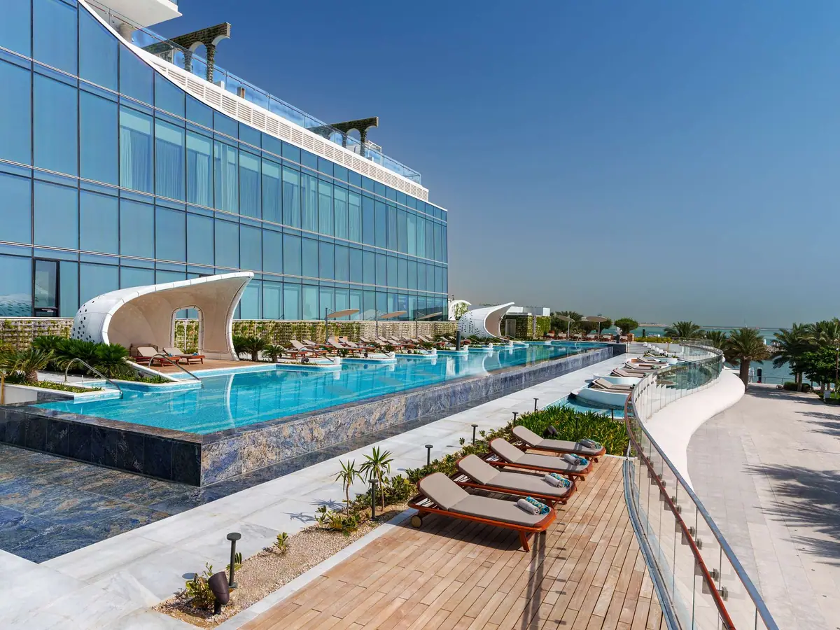 A very long pool lined with loungers beside a waterfront promenade