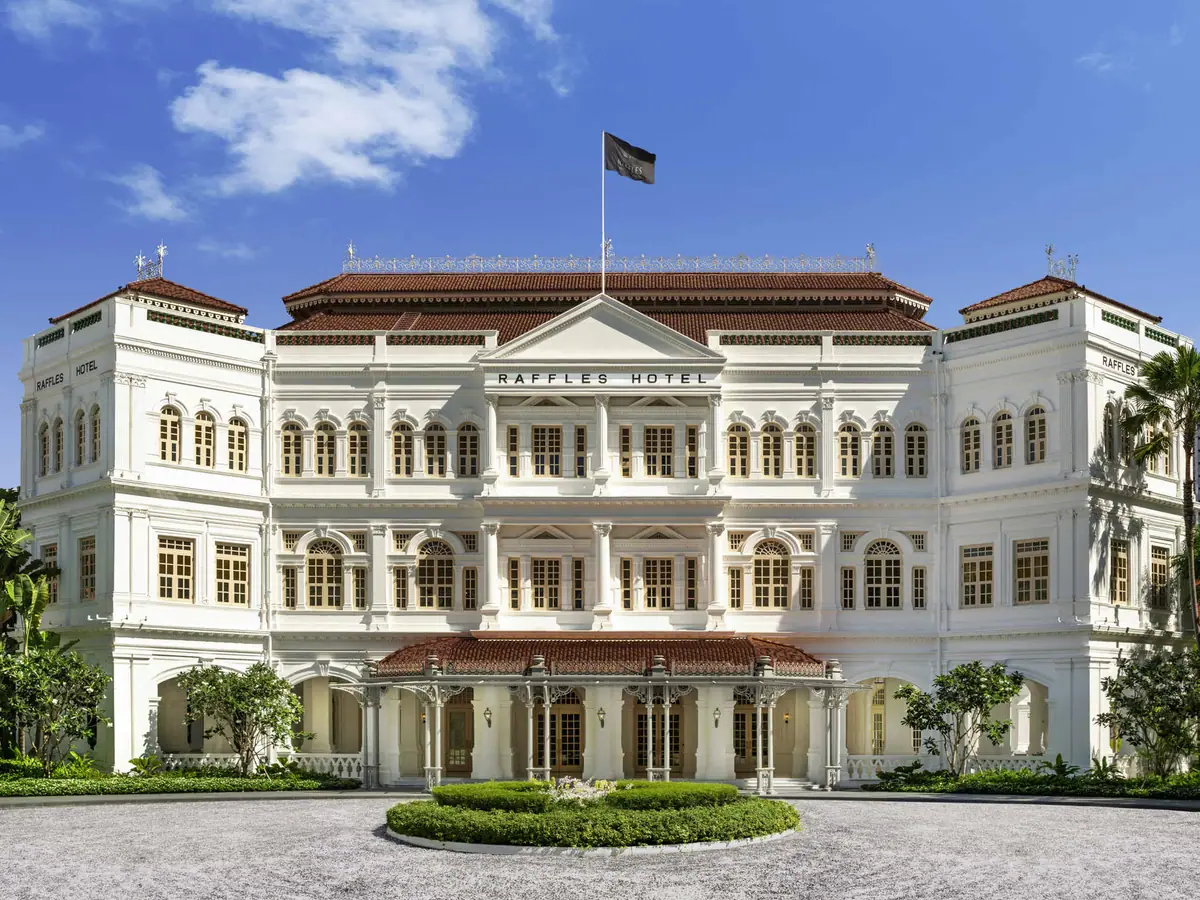 A white colonial building with arched verandas behind palms and a flag