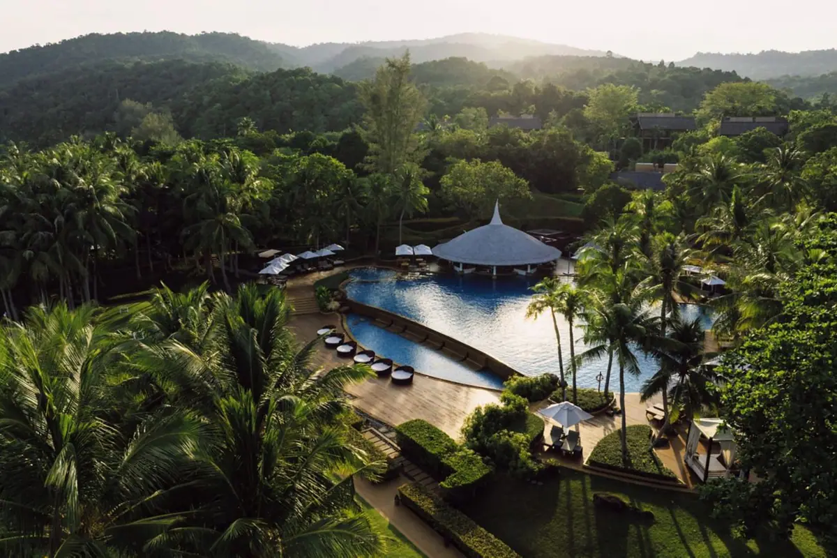 An aerial view of low roofs and a pool among palms above a bay