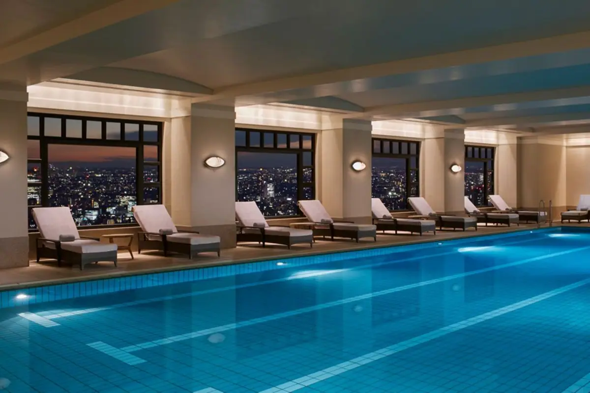 An indoor pool with loungers along one side and a wall of glass over a lit city