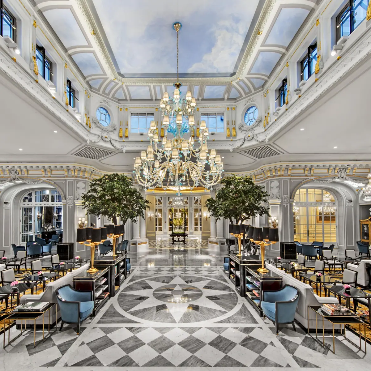 A high ceilinged hall with a chandelier, trees in pots and a chequered floor