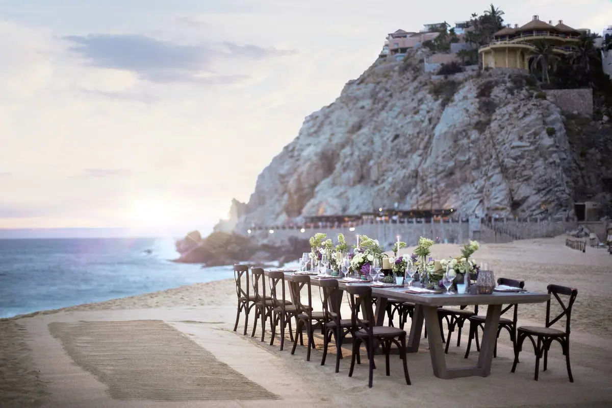 A long table laid for dinner on a beach at sunset with a rock headland behind