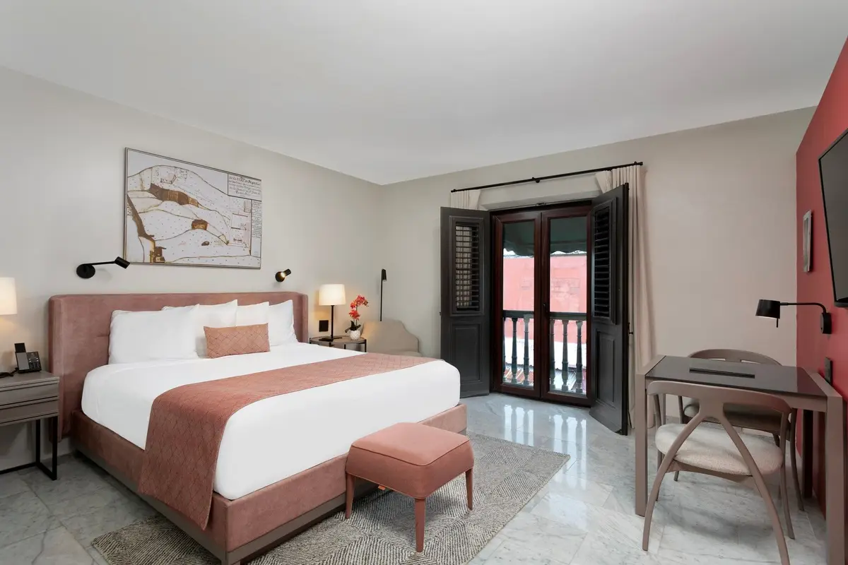 Guest room with a pink upholstered bed, a writing desk and shuttered doors to a small balcony