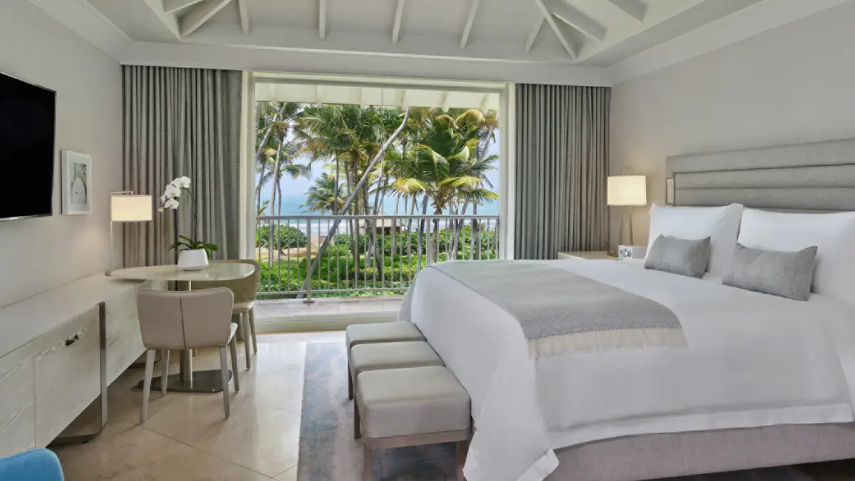 Guest room with a pale upholstered bed and glass doors onto a balcony over the palms