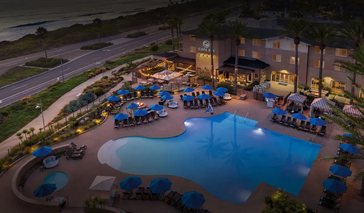 An aerial view at dusk of a low resort wrapped around a lit pool beside a coast road