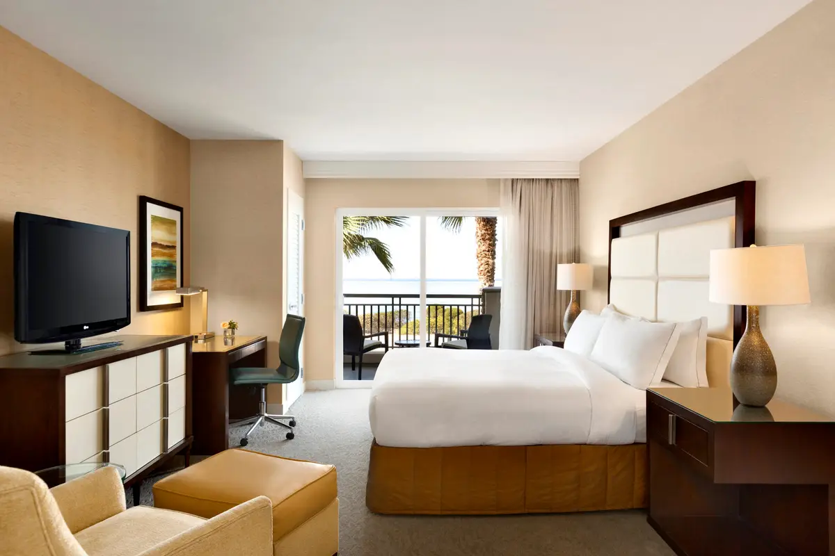 A guest room with a pale upholstered headboard, a desk and doors onto a balcony