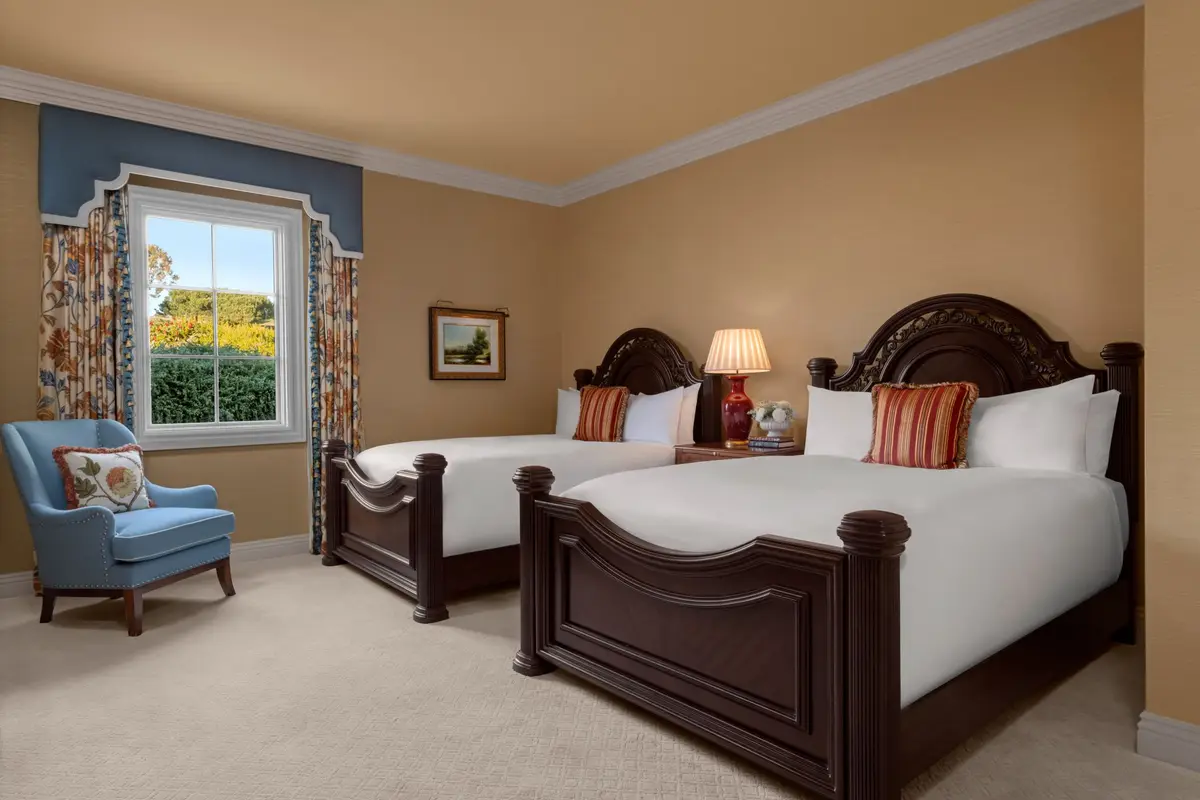 A guest room with two queen beds, a carved dark wood headboard and a window onto the gardens