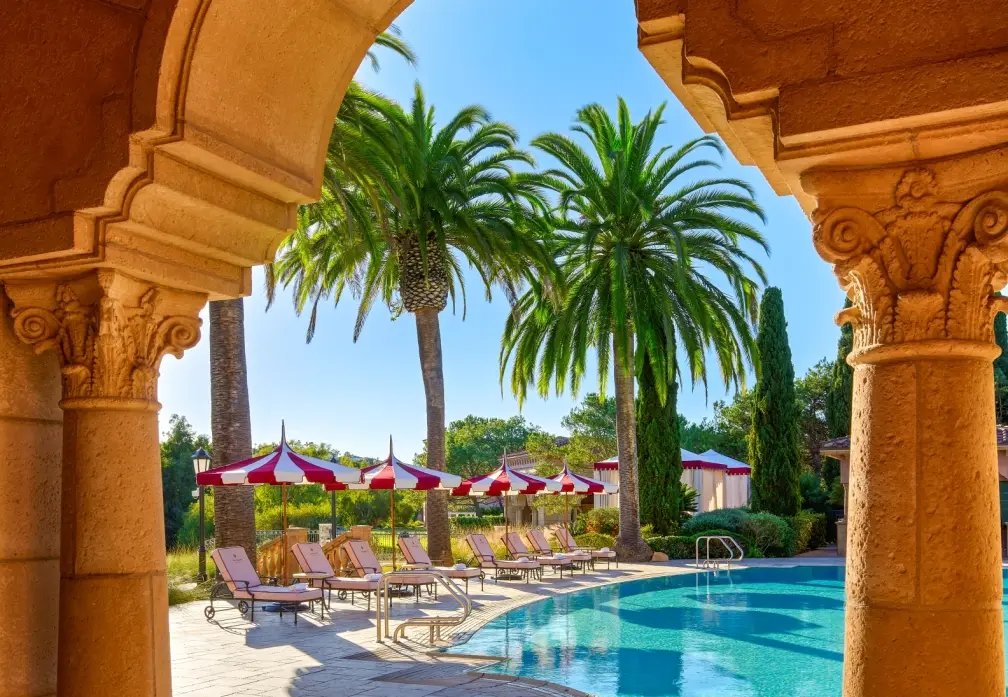 A resort pool lined with palms and striped umbrellas seen through a carved stone arch