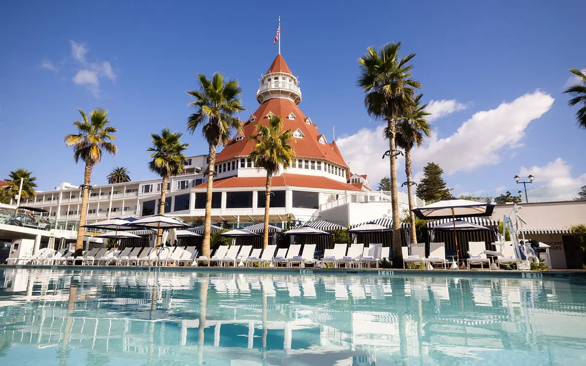 A pool lined with sun loungers and palms with the red roofs of the hotel behind