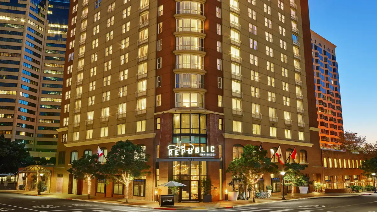A tall lit hotel tower on a downtown corner at dusk with street trees and flags below