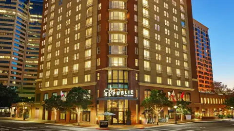 A tall lit hotel tower on a downtown corner at dusk with street trees and flags below