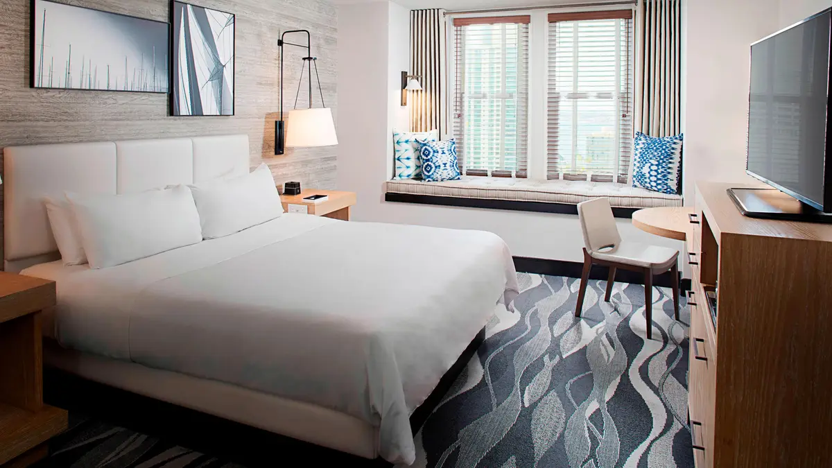 A guest room with a white bed, patterned grey carpet and a window seat looking over the city