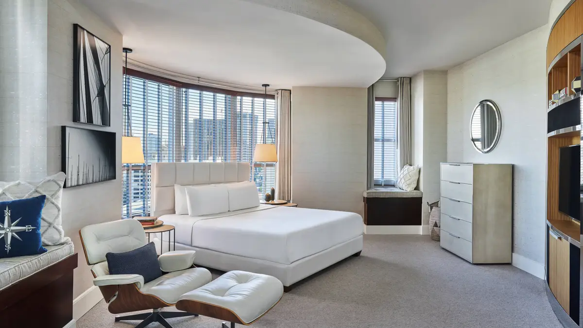 A curved corner suite bedroom with a low white bed, ottoman and wraparound windows